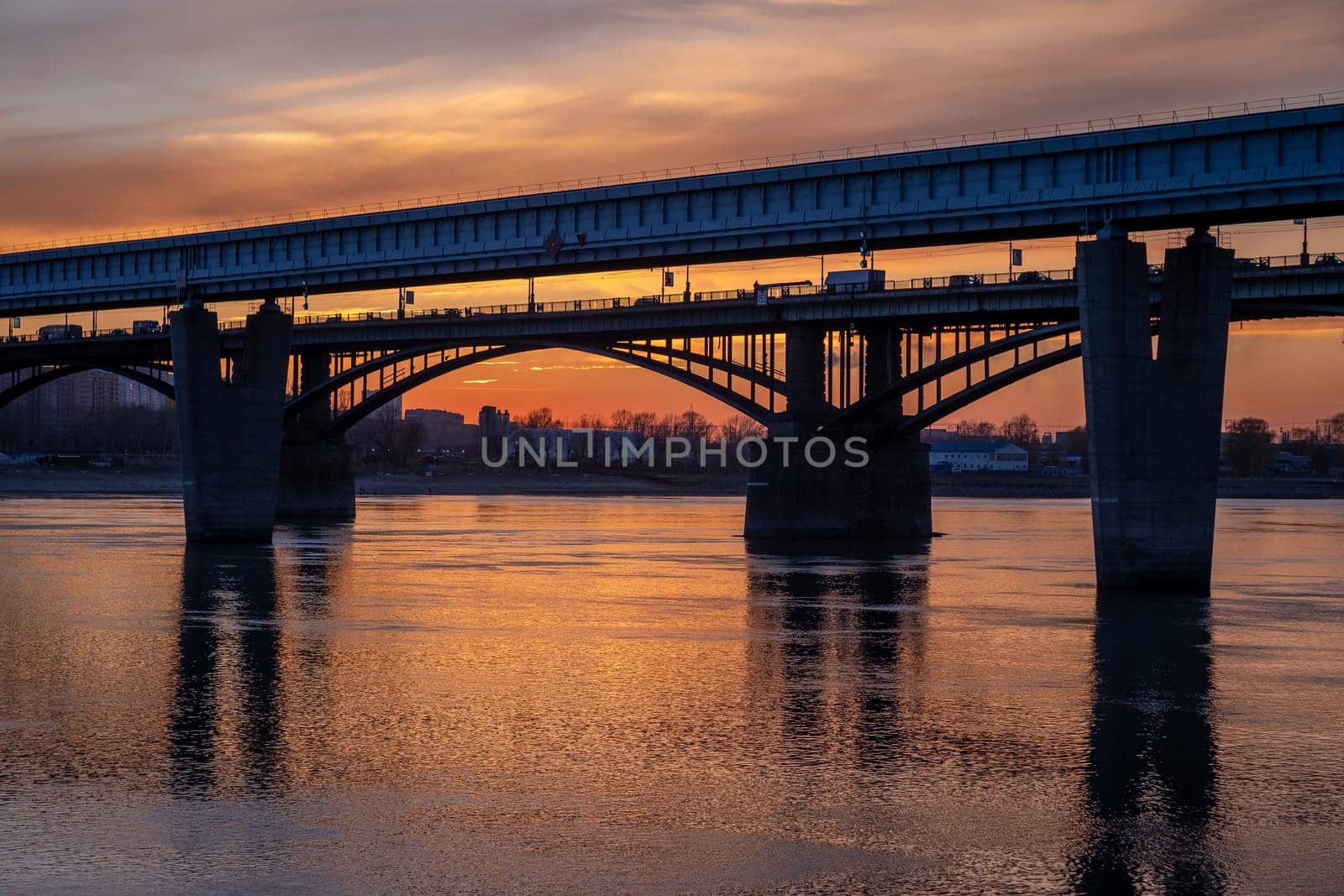 Beautiful view of the bridge over which cars drive at sunset. A river flows under the bridge, reflecting the sunset rays. Bridge in the city of Novosibirsk, Russia