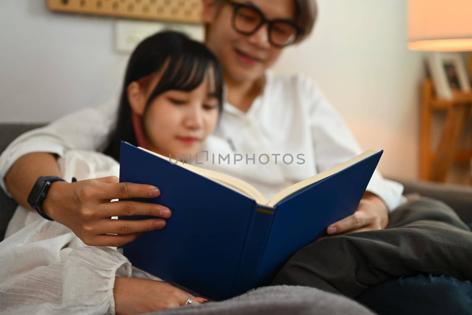 Loving young Asian couple reading book and relaxing together on sofa in living room.
