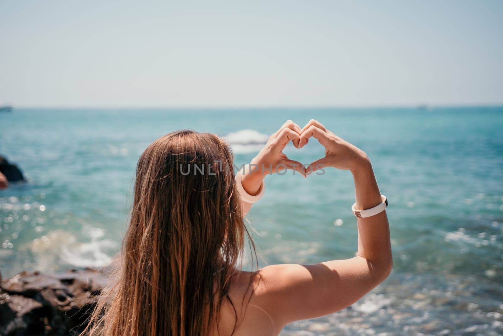 Woman makes heart with hands on beach. Young woman with long hair in white swimsuit and boho style braclets practicing by sea ocean. Concept of longing daydreaming love peace contemplation vacations by panophotograph