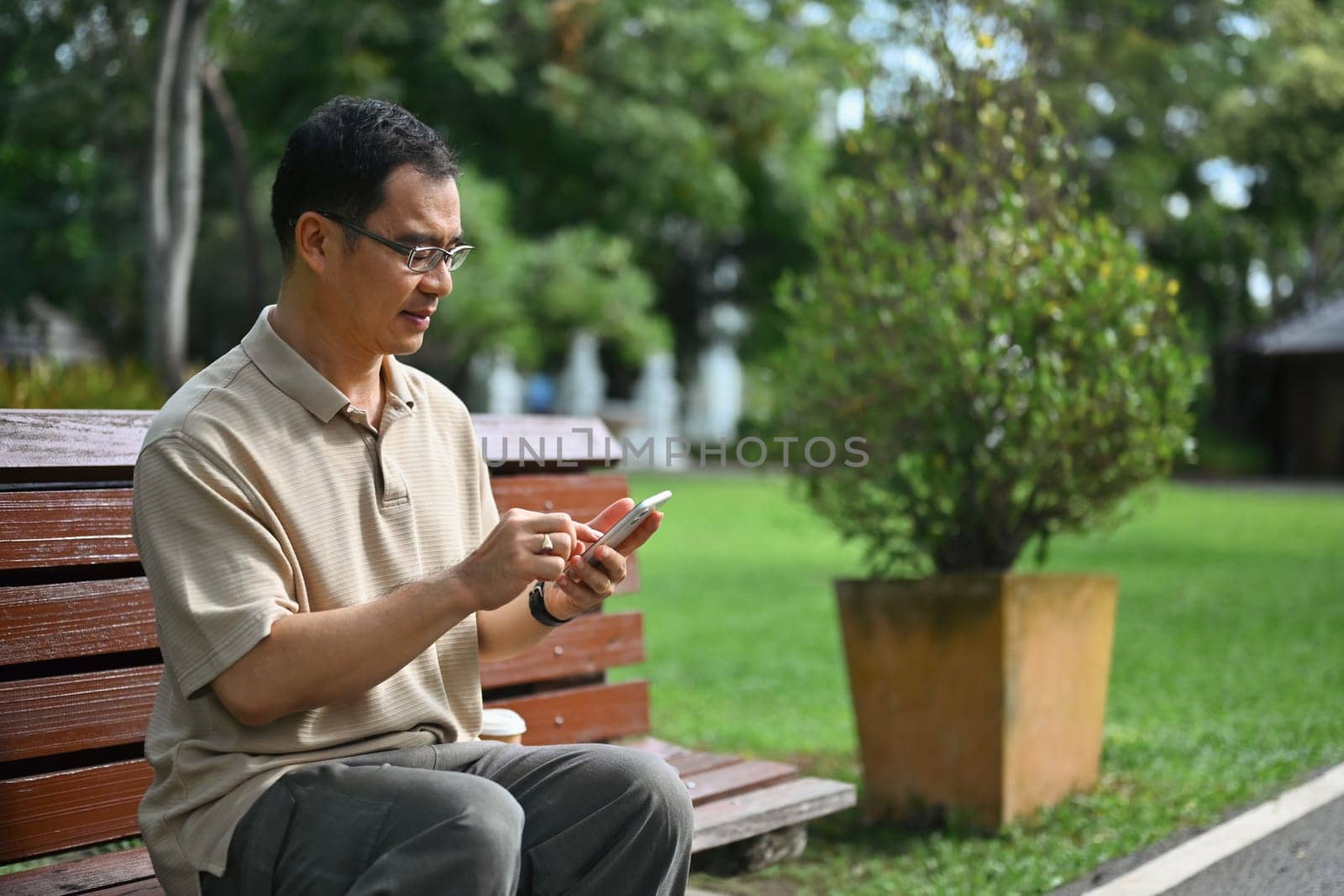 Happy relaxed middle aged man using mobile phone sitting on a wooden bench at the park by prathanchorruangsak