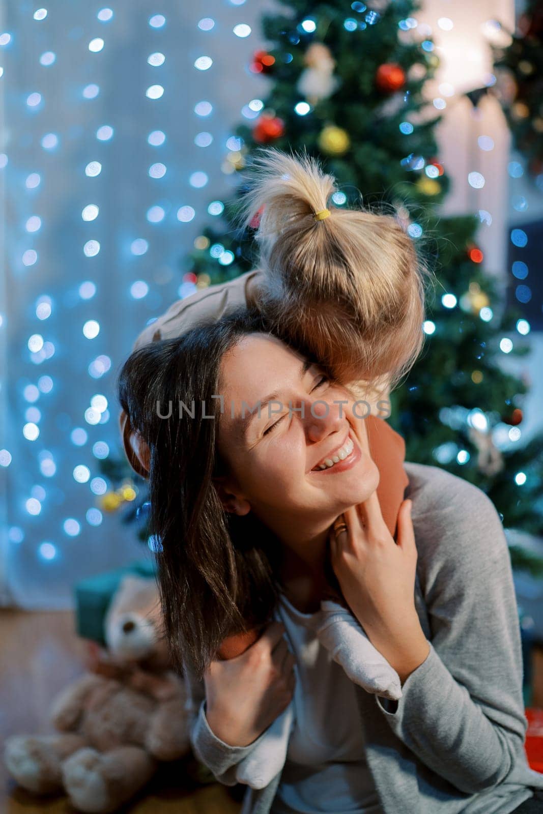 Little girl hugged her smiling mother sitting on her shoulders near the Christmas tree. High quality photo