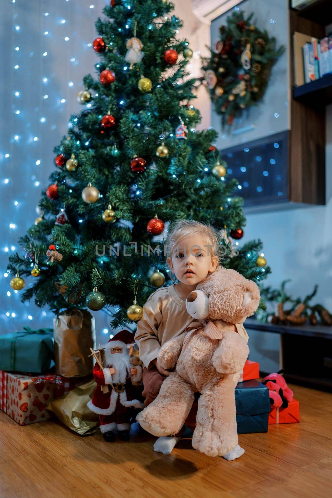 Little girl sits under a Christmas tree hugging a teddy bear by Nadtochiy