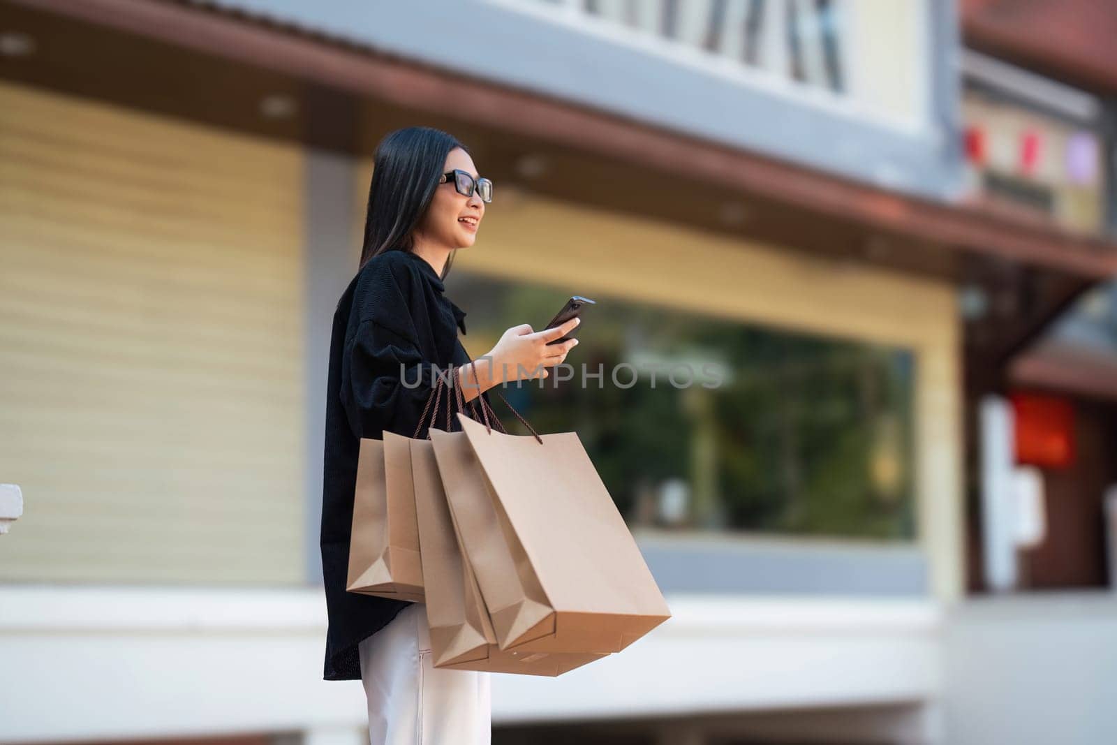 Woman using smartphone holding Black Friday shopping bag while standing on the side with the mall background.