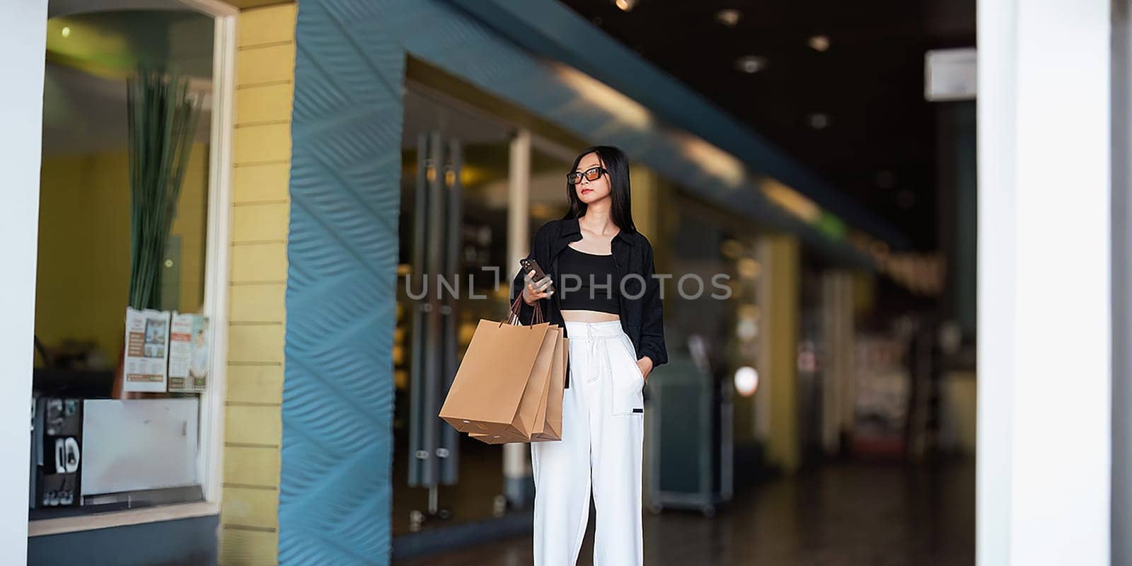Woman using smartphone holding Black Friday shopping bag while standing on the side with the mall background by nateemee