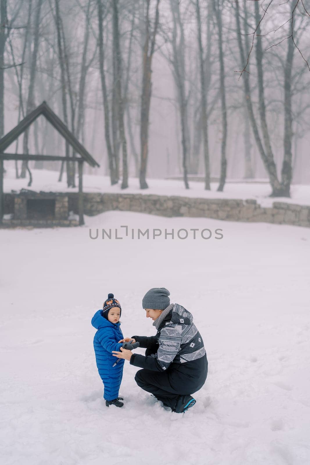 Mom puts on mittens to a little girl squatting in a snowy forest. High quality photo