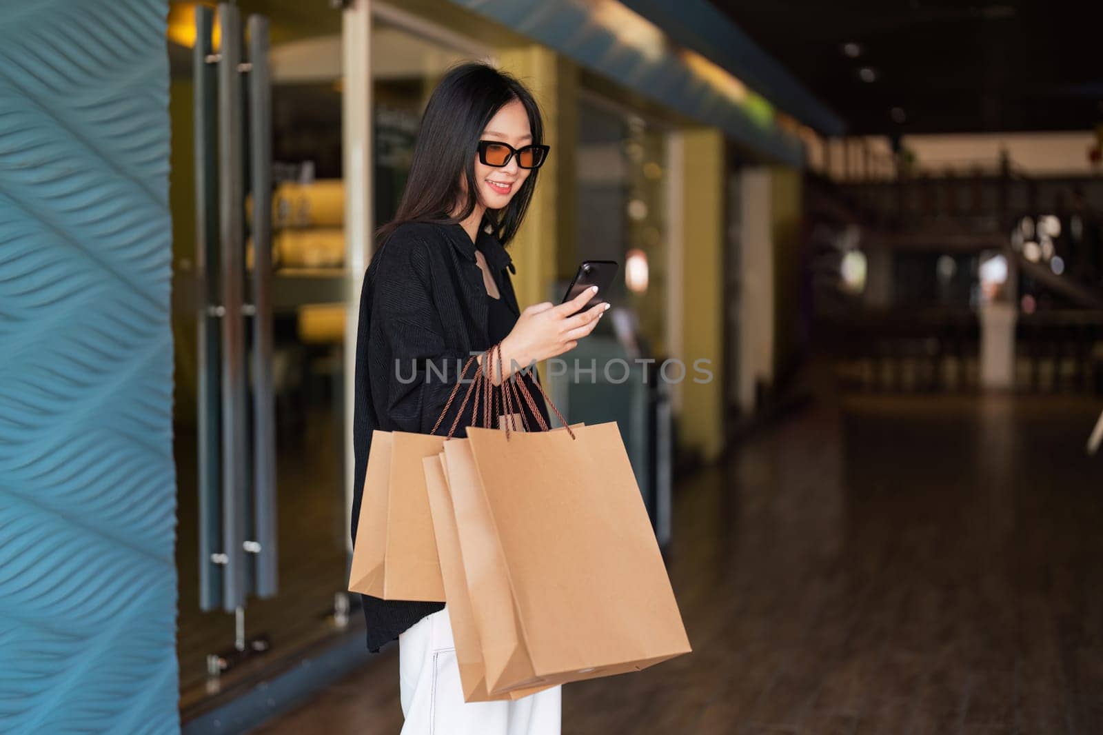 Woman using smartphone holding Black Friday shopping bag while standing on the side with the mall background by nateemee