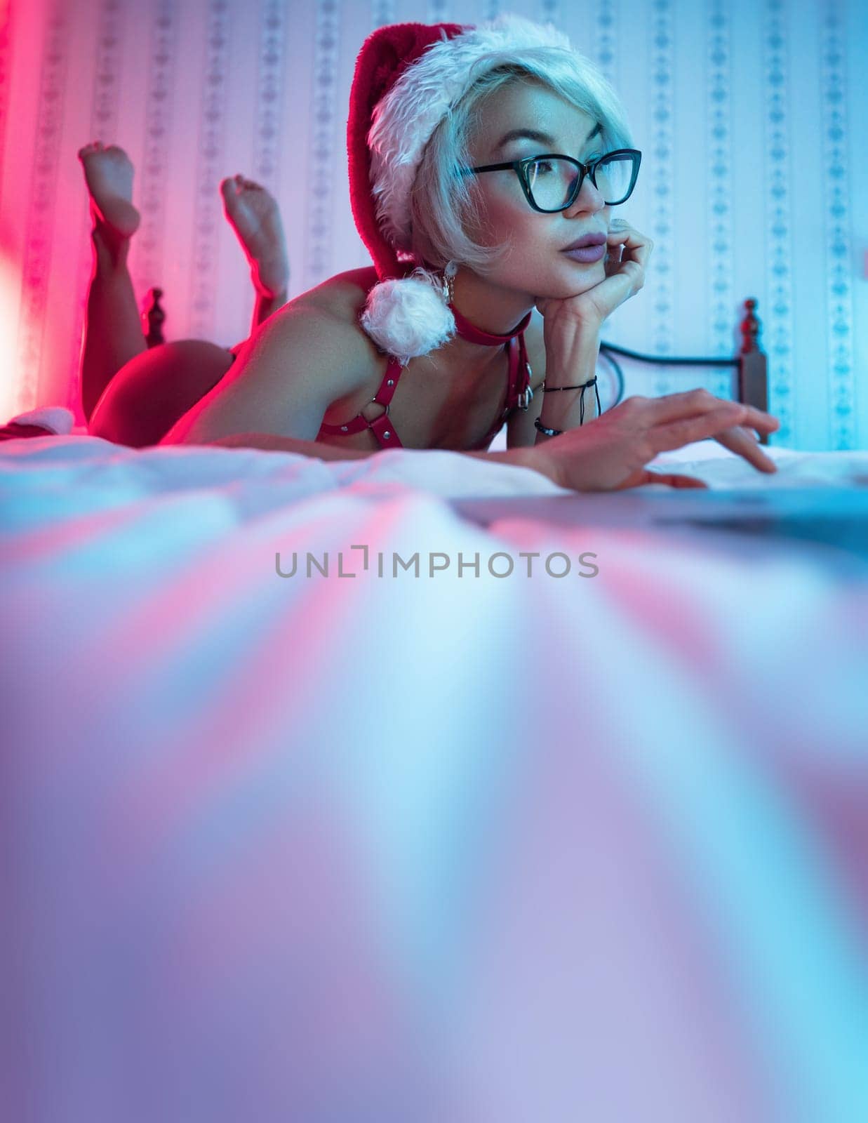 Sexy girl in lingerie wearing a Santa Claus hat poses sexily on a bed with a laptop on Christmas Day in neon lights