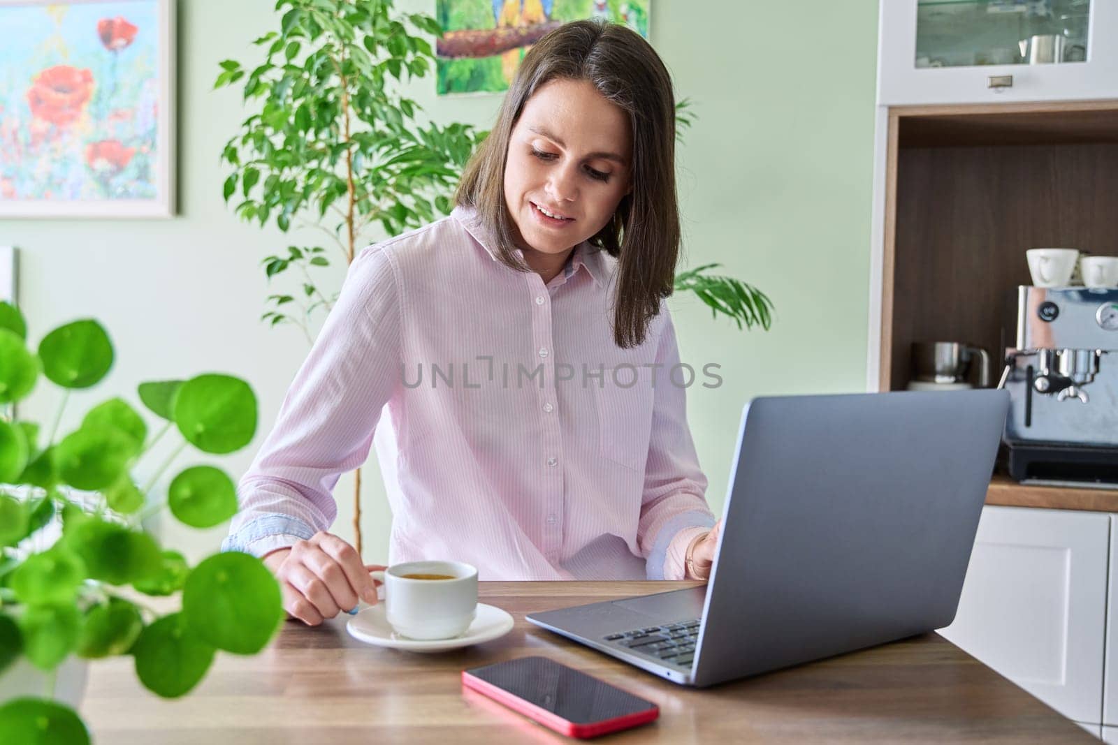 Young woman sitting at home in kitchen using laptop, drinking morning coffee. Lifestyle, freelancing, technology and work leisure, 25-30 year old people concept