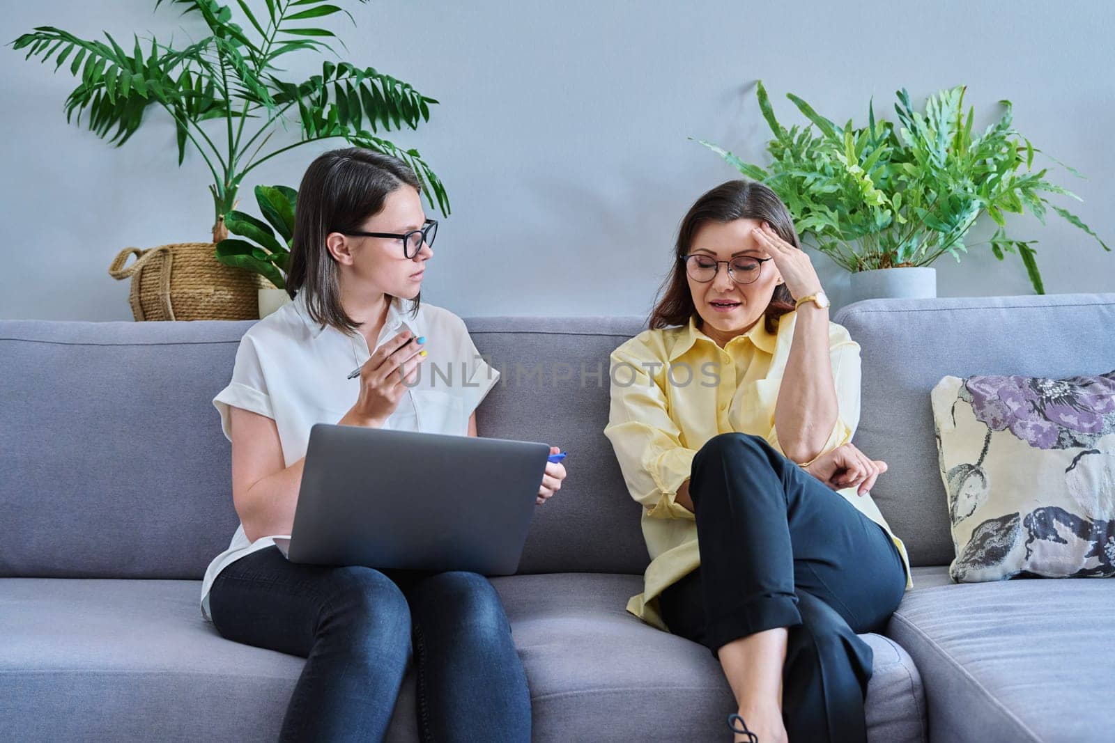 Female psychologist and middle aged woman patient sitting together on couch in office by VH-studio