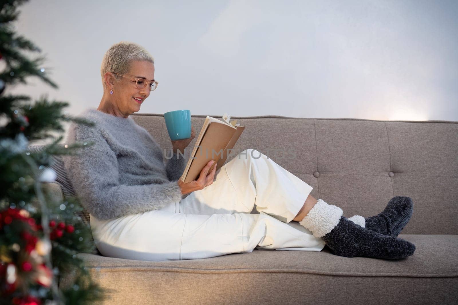 Smiling elderly woman with a cup of tea and a book relax in side of a decorated Christmas tree in living room.