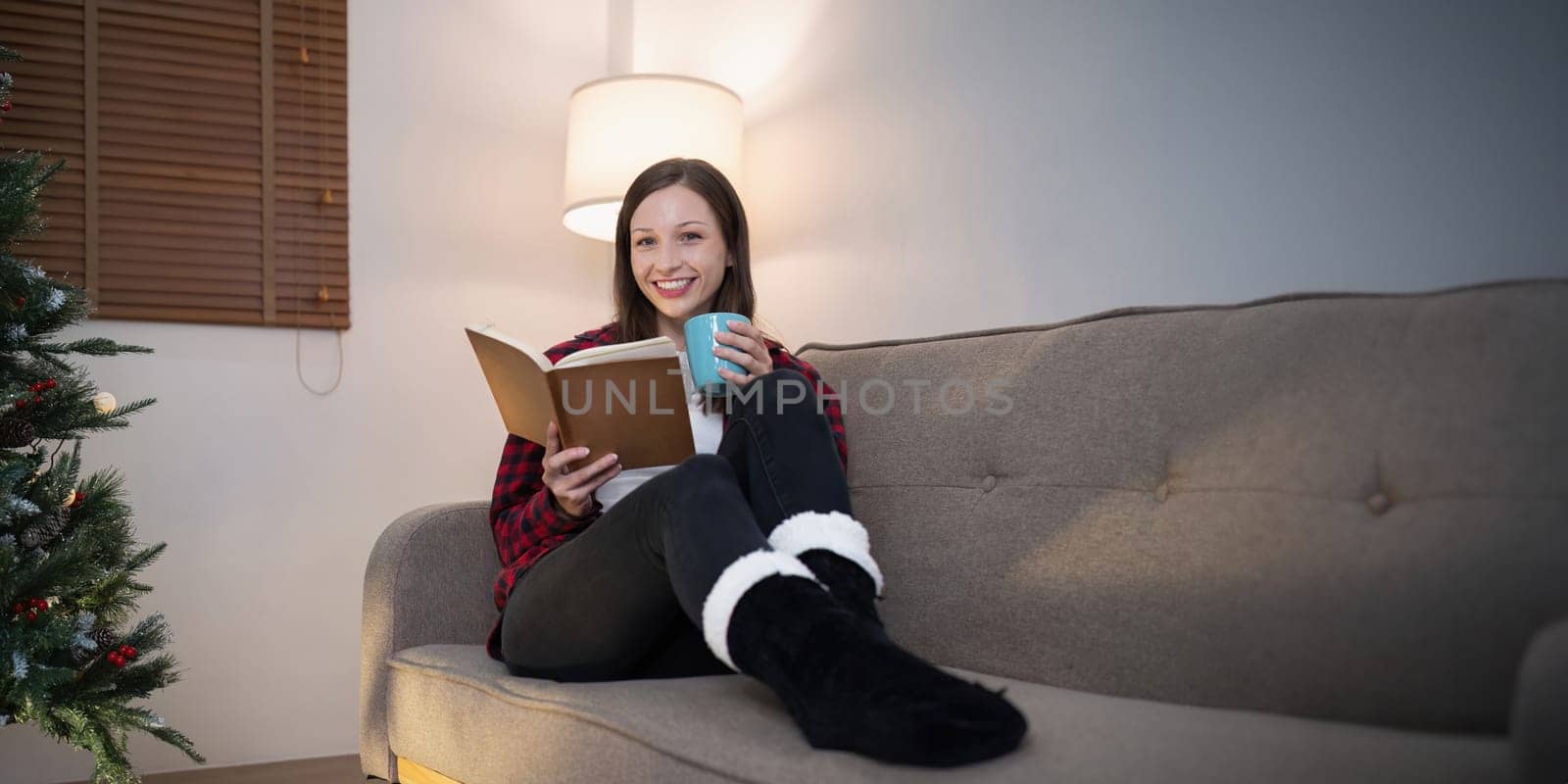 Young cute woman holding cup coffee or tea and read interest book. Concept of Christmas atmosphere at home by nateemee