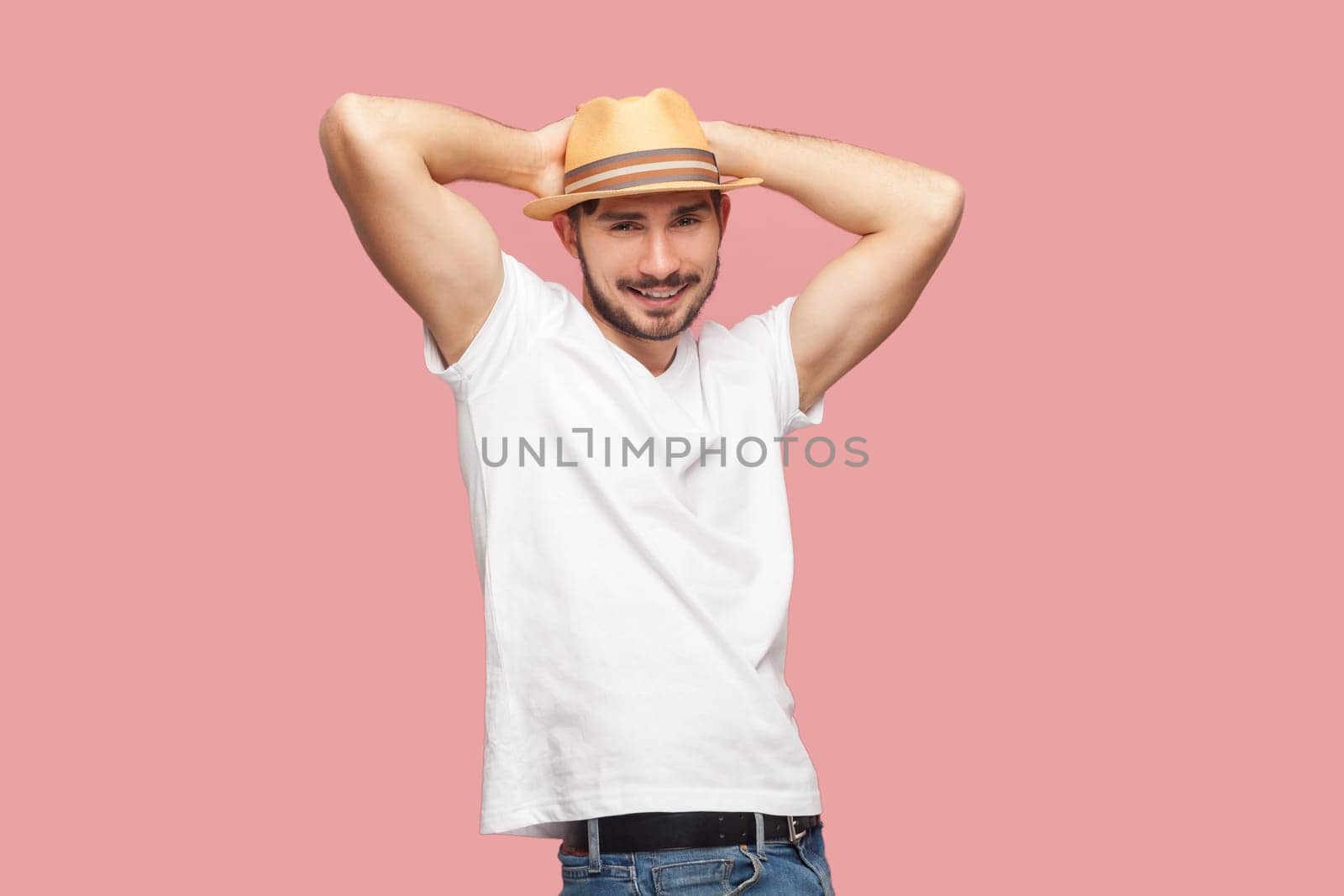 Portrait of smiling attractive handsome bearded hipster man in white T- shirt and casual hat standing with hands behind head, looking at camera. Indoor studio shot isolated on pink background.