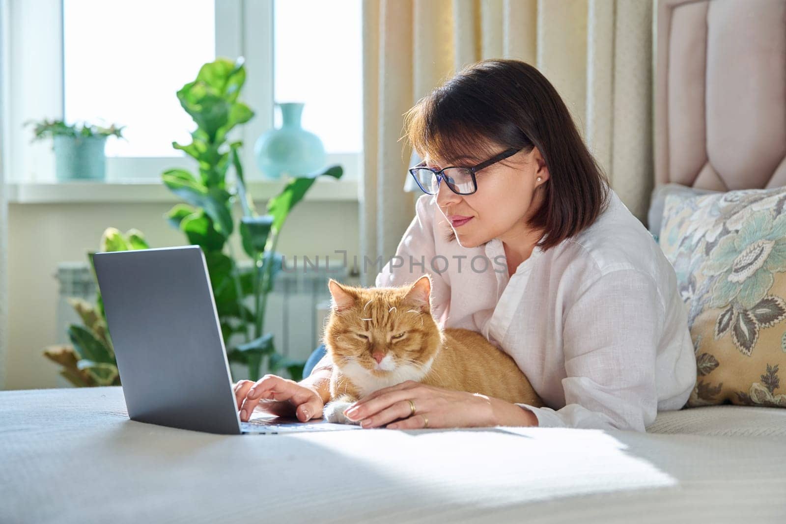 Middle aged woman at home on couch with laptop and cat. Mature 40s female looking at camera, on bed with pet. Work at home, technology, leisure, freelancing, lifestyle, people and animals concept