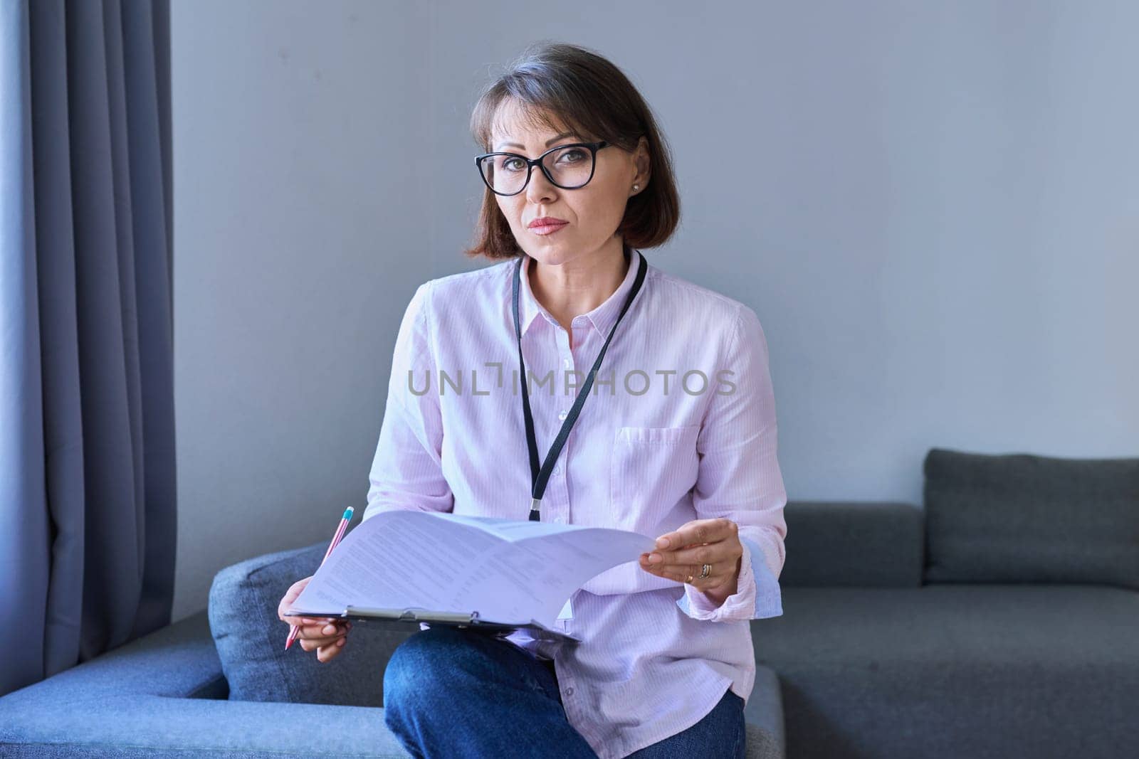 Business 40s woman with clipboard sitting on couch in office. Middle aged female advisor, accountant, agent, assistant, workplace
