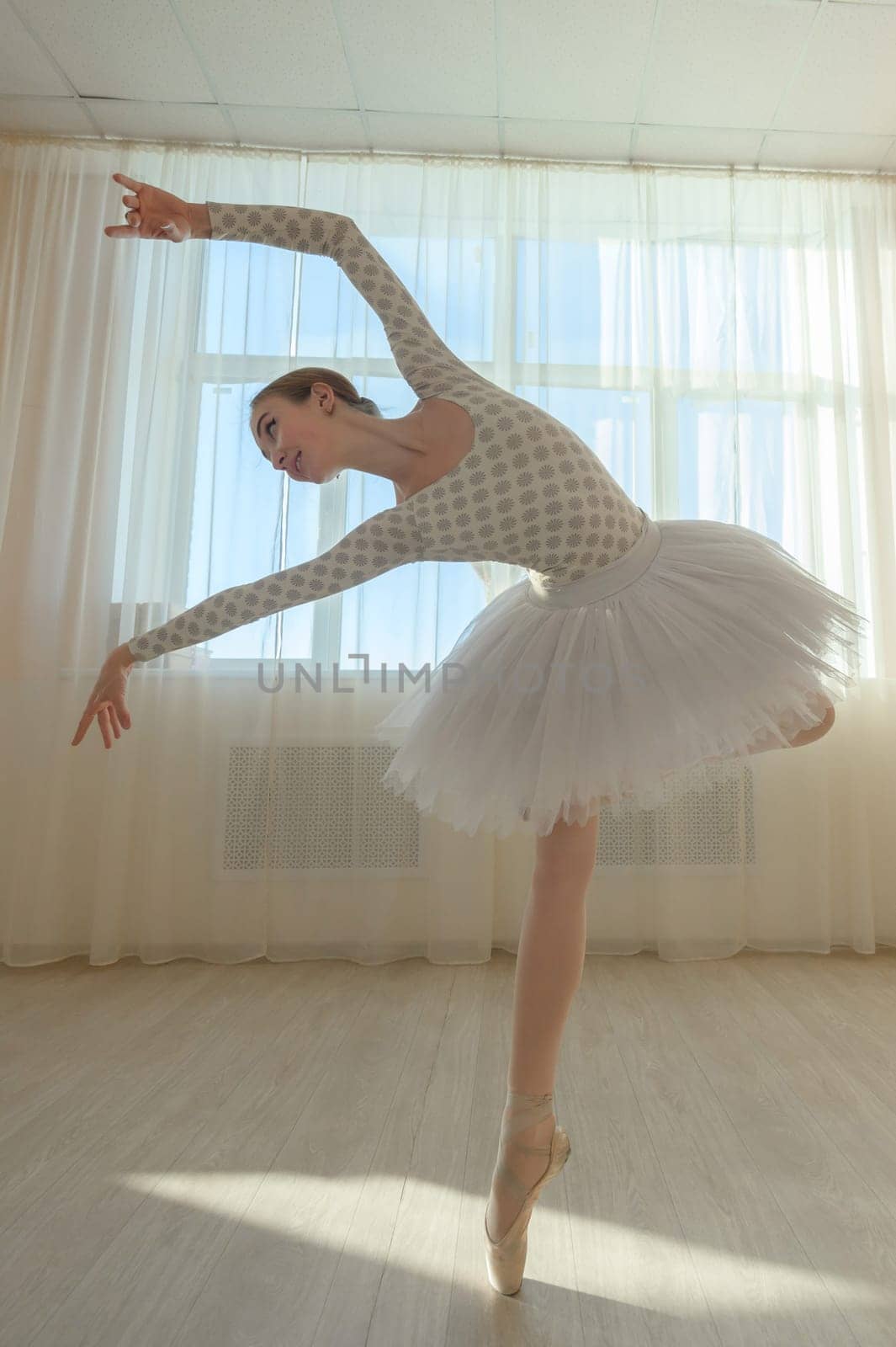 Beautiful graceful ballerina is practicing in the hall against the background of a window. Slender woman in tutu and pointe shoes in a dance class