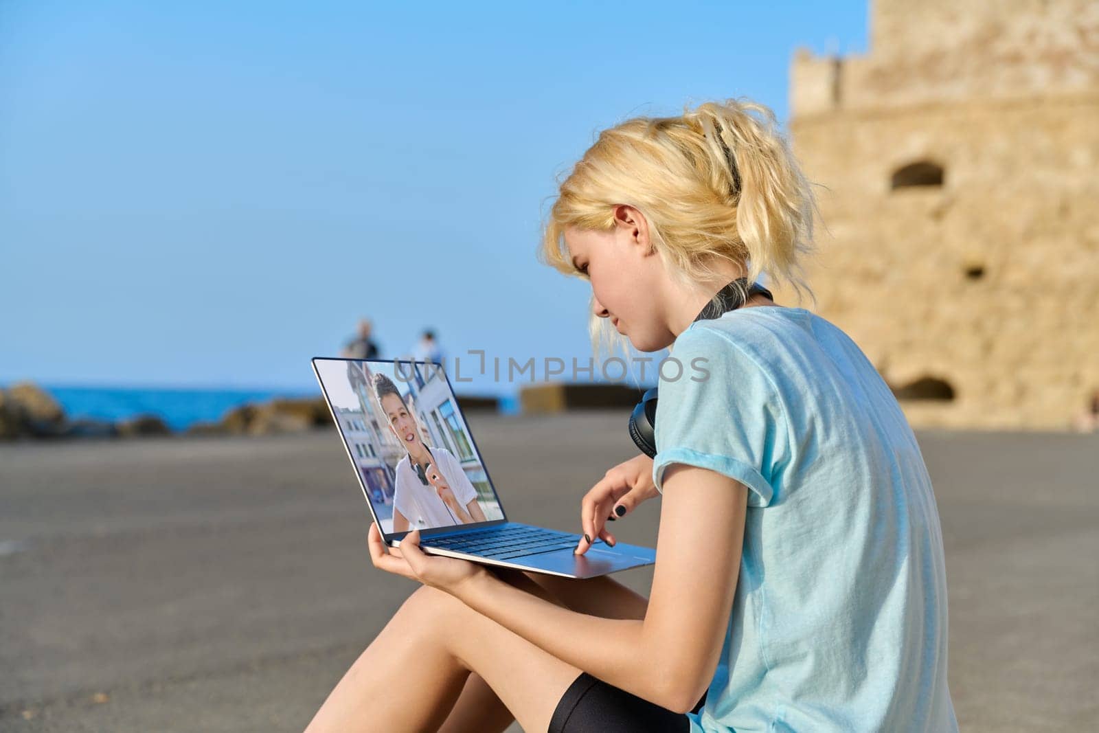Video call on laptop, teenage friends talking online. Web chat of teenage girl sitting on seashore in European tourist destination and guy. Technology, tourism, friendship, lifestyle, young people concept