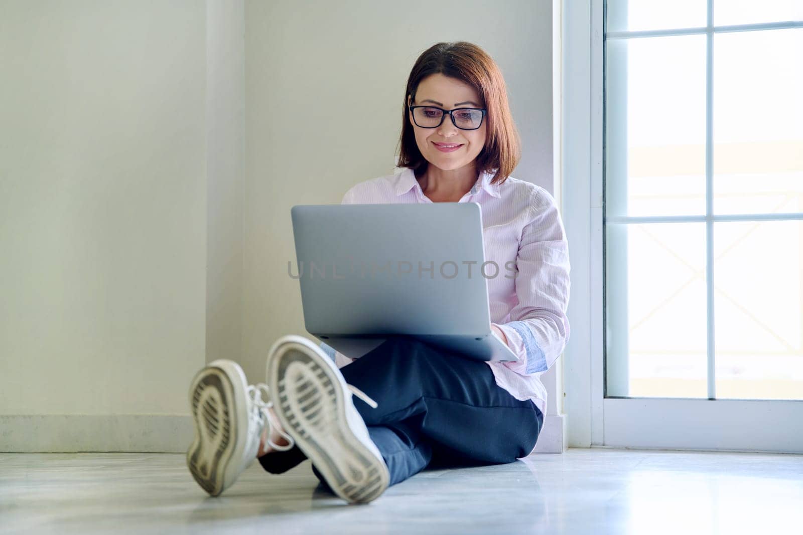 Middle aged woman sitting on floor using laptop, window wall background. Freelancing, remote work, leisure, technology, lifestyle, people concept