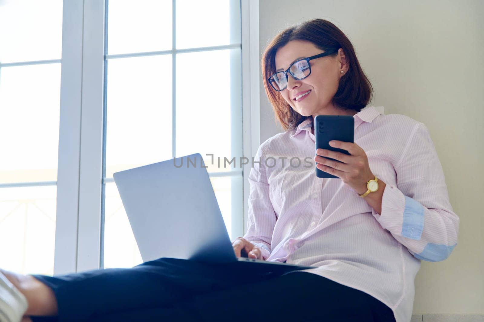 Business mature woman sitting on floor at home using laptop and smartphone. Remote business, work, technology, 40s people concept