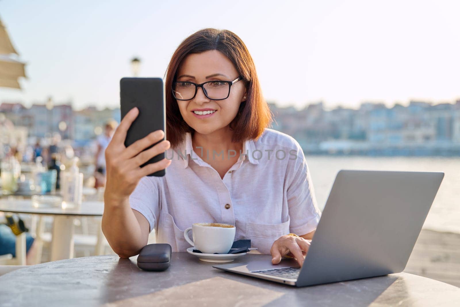 Smiling successful middle aged business woman sitting in outdoor cafe with cup of coffee using laptop and smart phone. Business, technology, freelance, remote work, 40s people concept