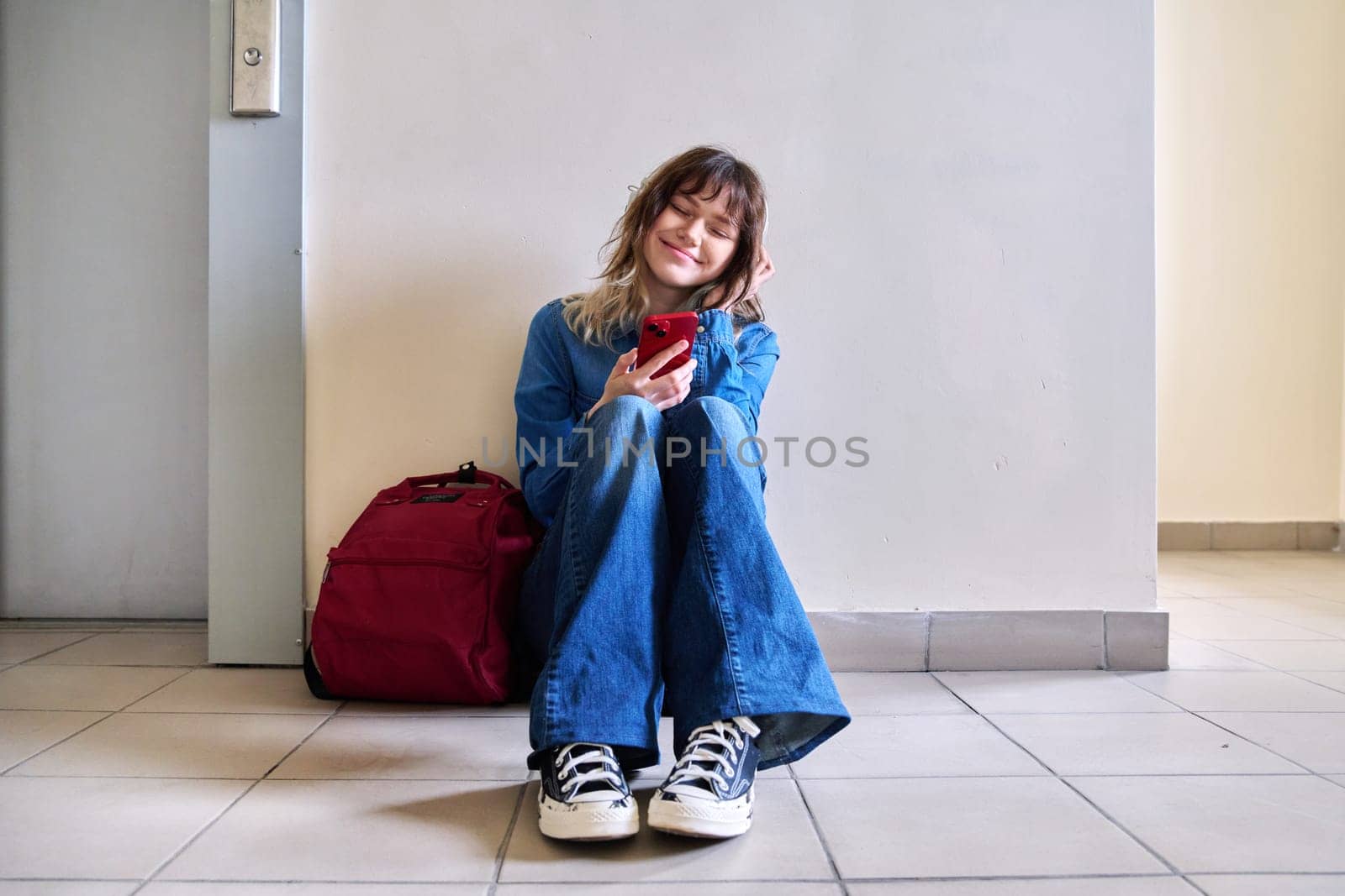 Happy teenage girl in headphones enjoying music with closed eyes. Young female student with backpack sitting on floor near elevator. Music, joy, happiness, youth, young people concept