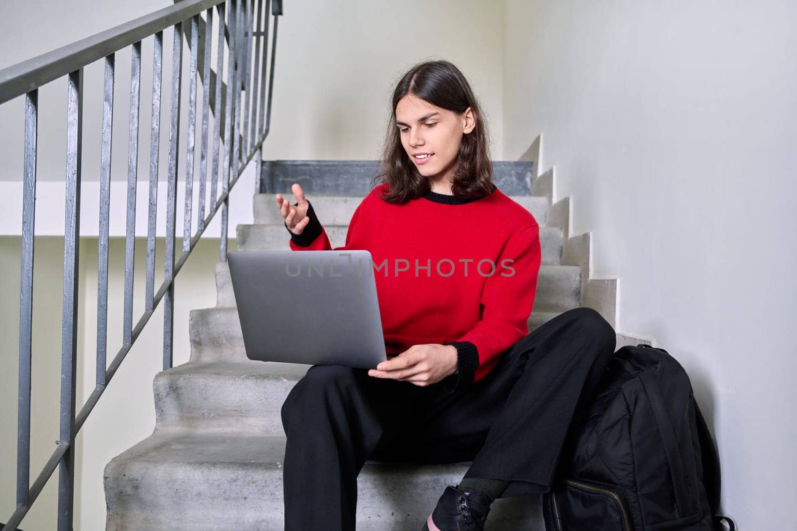 Portrait of handsome student guy with backpack on steps inside building using laptop, talking on video chat. Youth, college, university, education, technology, lifestyle, young people concept