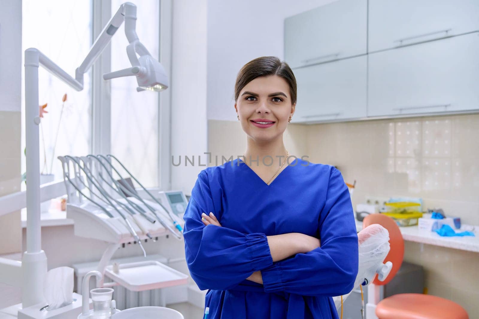 Portrait of confident female dentist in office looking at camera. Young female doctor in blue uniform with crossed arms, copy space. Dentistry, medicine, health care, profession, stomatology concept