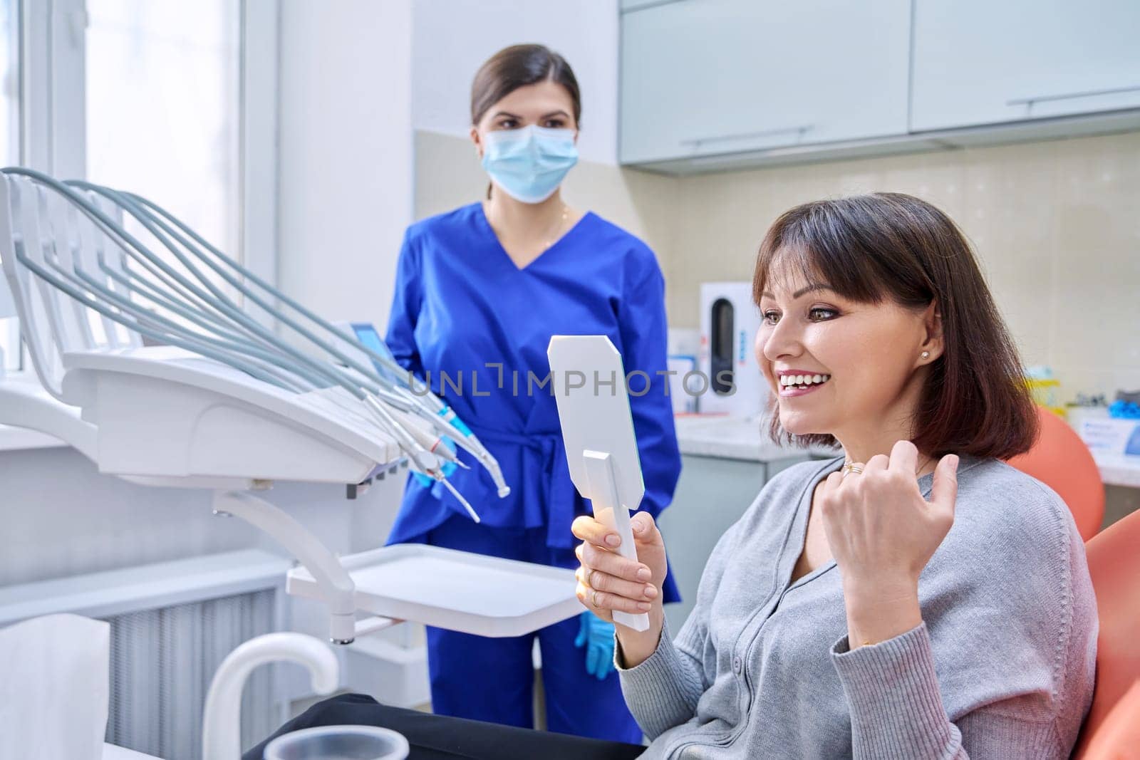 Dentist's office, woman patient looking at her teeth in the mirror. Doctor dentist near the dental chair with tools. Treatment, dental care, prosthetics, orthodontics, dentistry concept