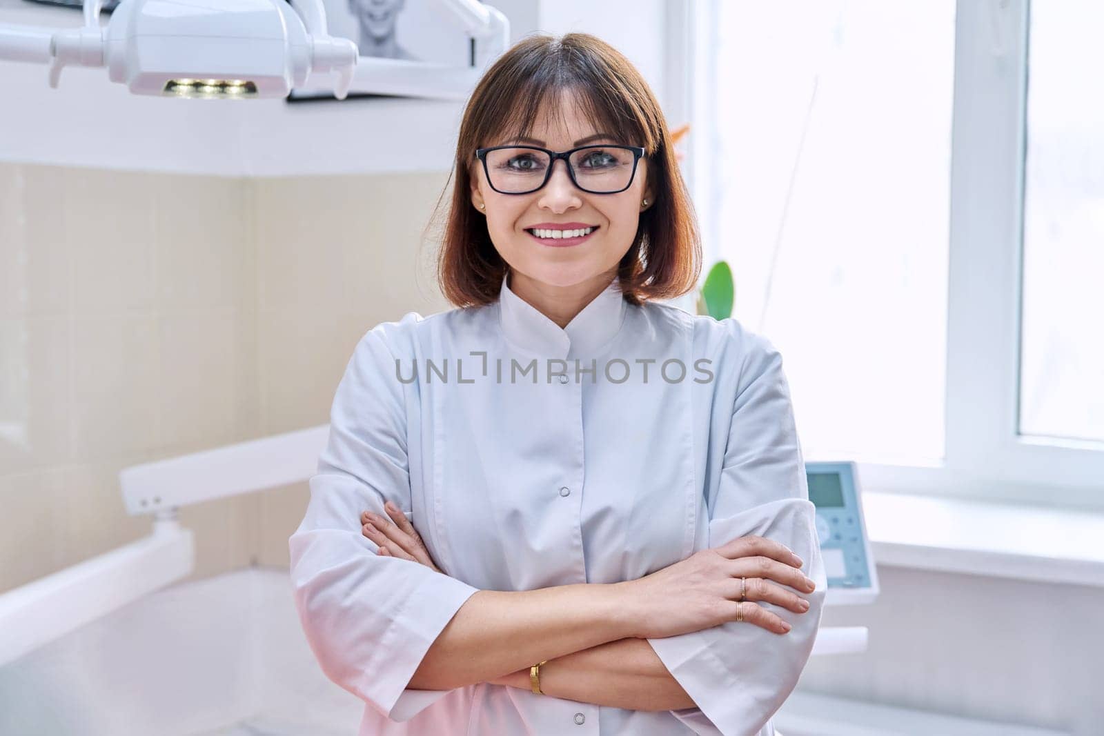Portrait of smiling mature female dentist looking at camera in office. Confident doctor with crossed arms in clinic. Dentistry, medicine, health care, profession, stomatology concept