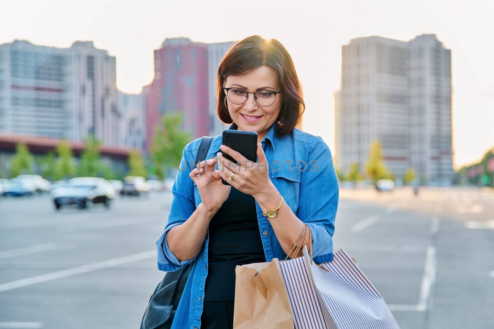 Middle aged woman using smartphone in the city. Beautiful confident successful female 40s age with shopping paper bags in mall parking, in sunset evening light
