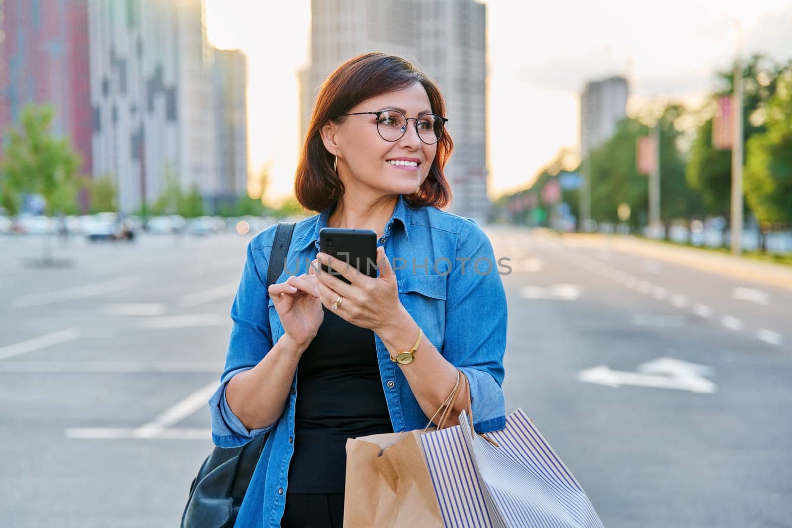Middle aged woman using smartphone in the city. Beautiful confident successful female 40s age with shopping paper bags in mall parking, in sunset evening light