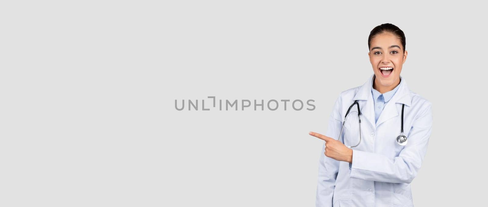 Shocked happy european millennial doctor in white coat, with open mouth, point finger at free space, isolated on gray studio background. Medical ad and offer, health care, sale surprise