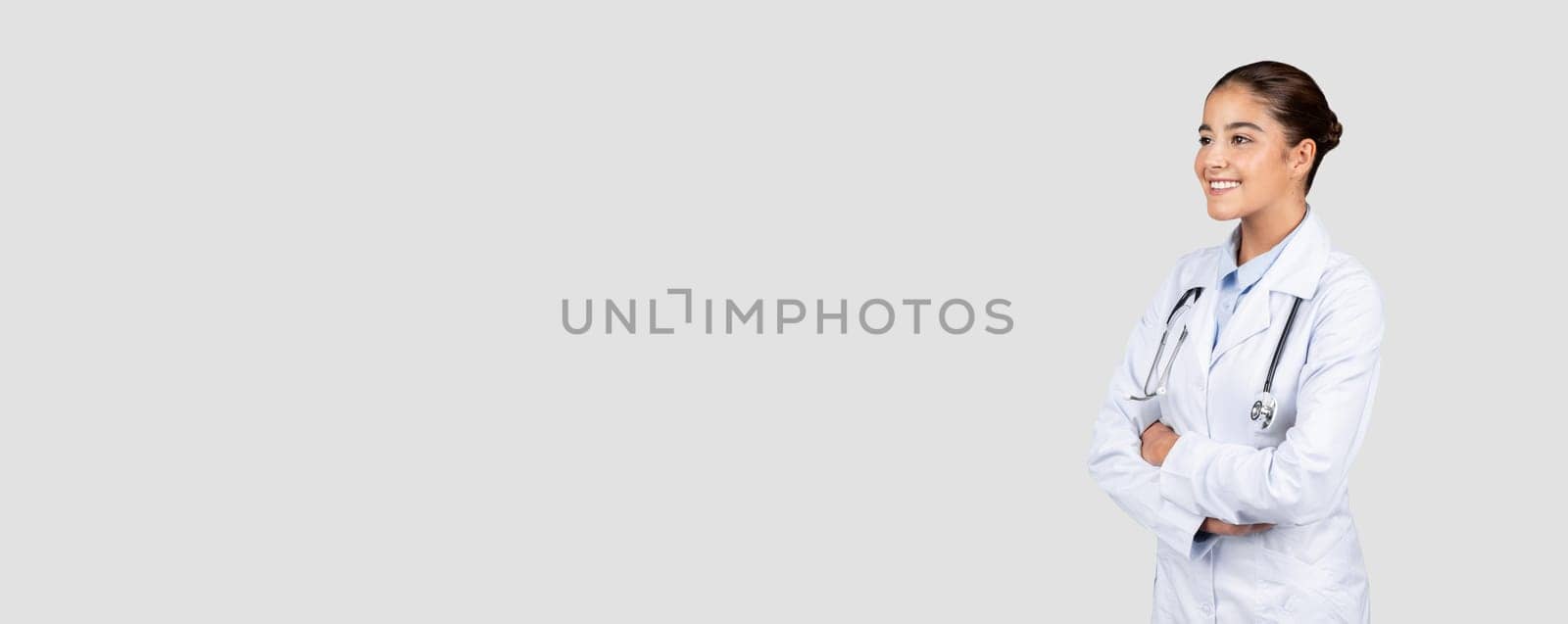 Confident smiling european millennial doctor in white coat, with stethoscope, look at free space, isolated on gray studio background. Medical ad and offer, health care, sale