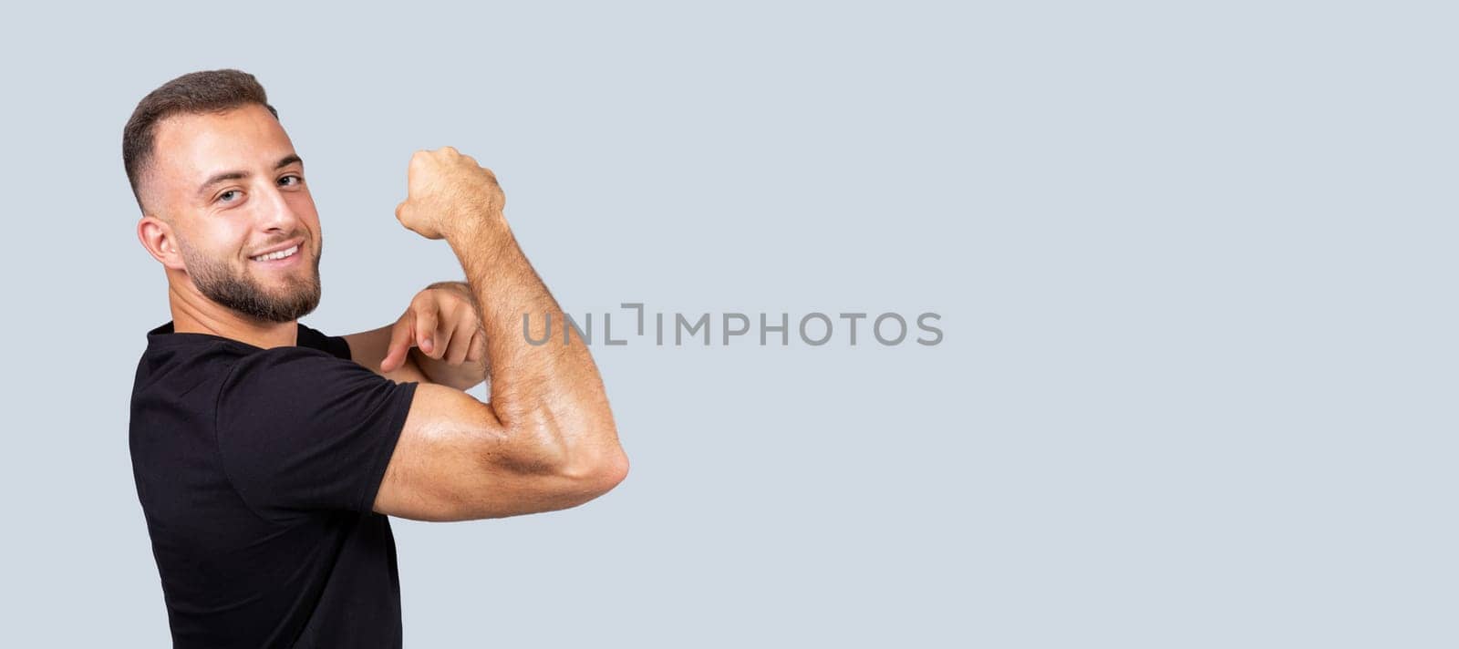 Cheerful confident strong millennial european man with beard in sportswear point finger at muscle biceps on hand, isolated on gray background. Body care, sport lifestyle and workout recommendation