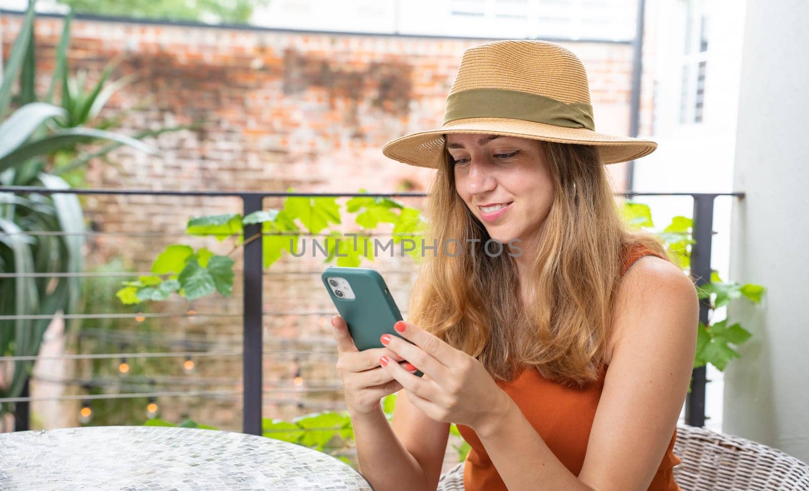 Happy attractive woman with hat using smartphone smiling on a terrace outside. High quality photo