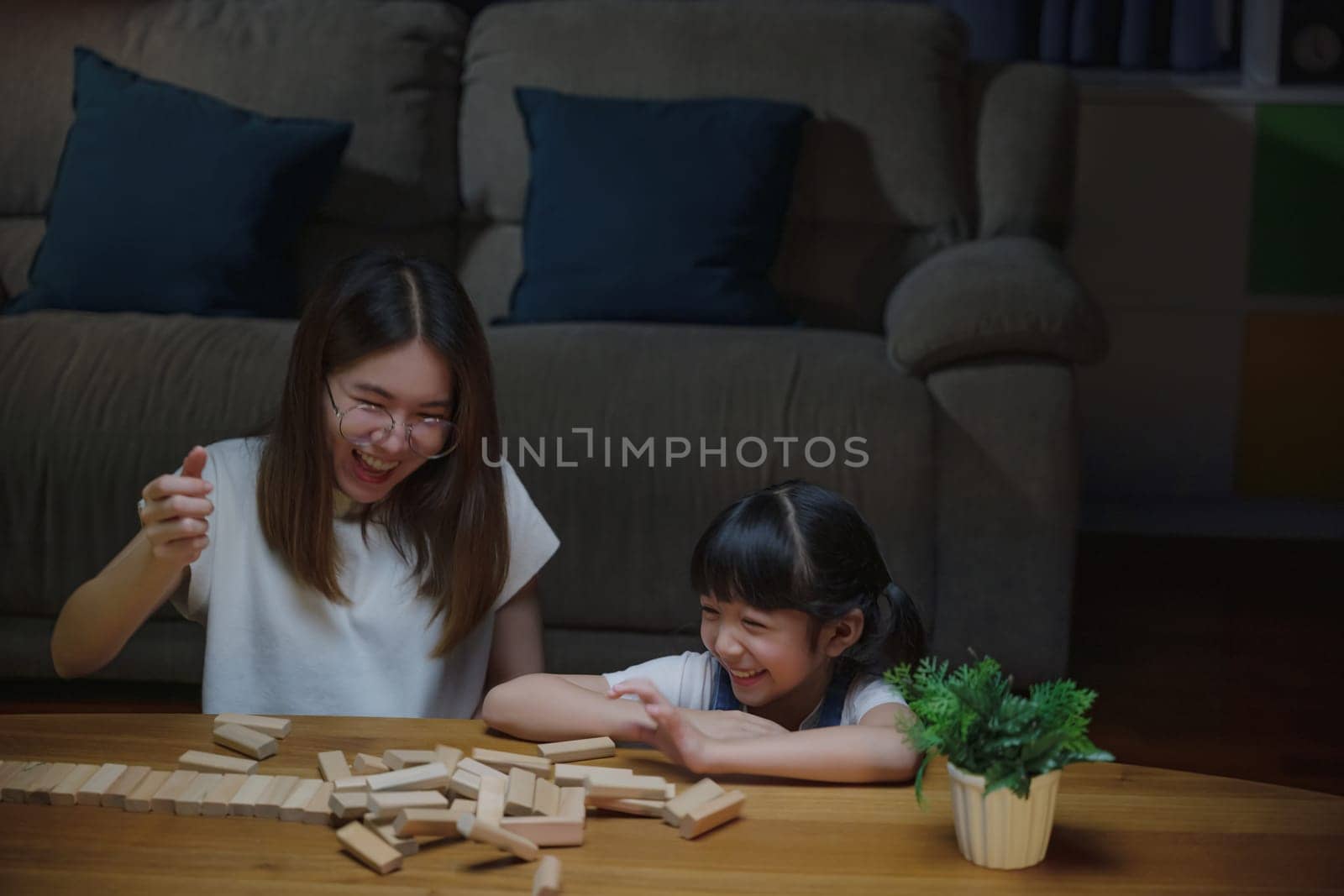 Asian young mother playing game in wood block with little daughter in home living room at night, Smiling woman help teach preschooler kid play build constructor tower of wooden blocks, family funny