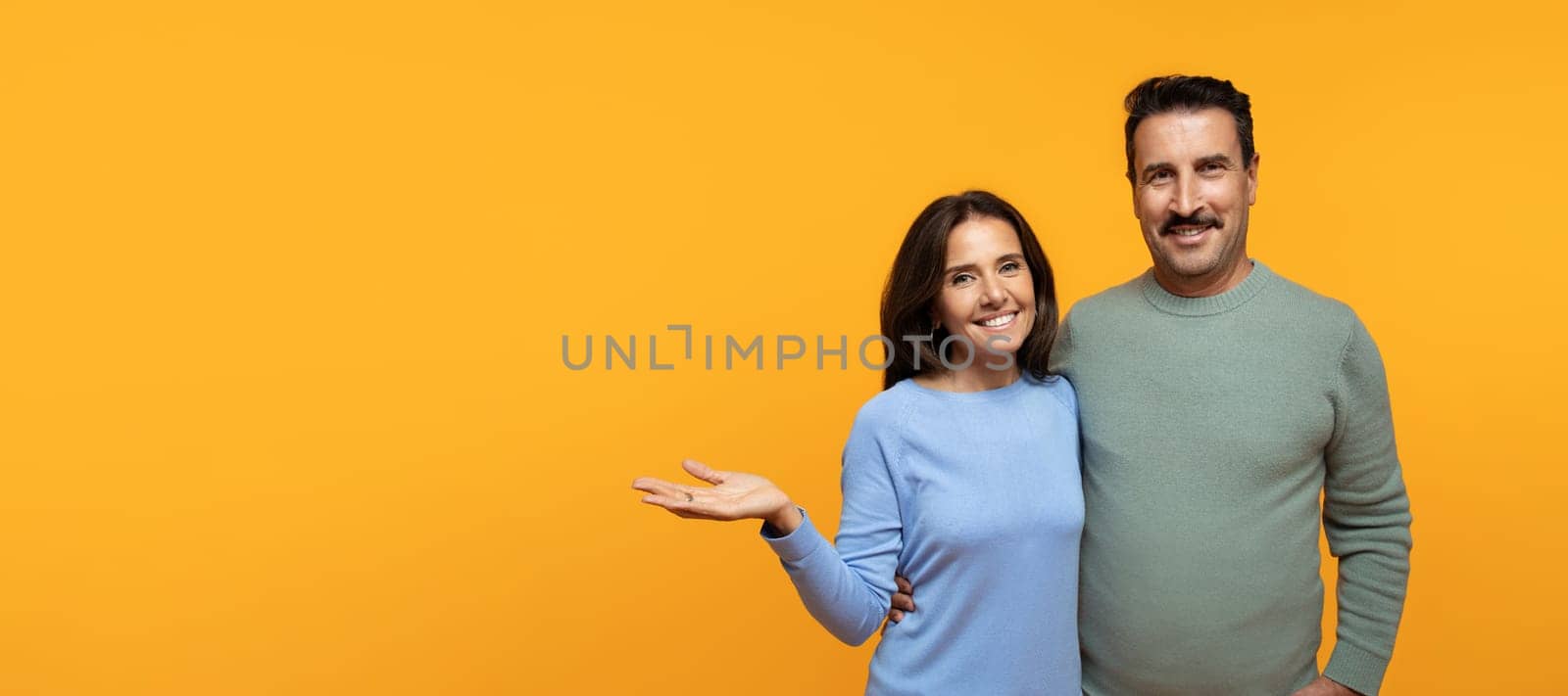 Smiling senior caucasian lady in casual hug man, hold on hand free space, isolated on orange studio background. Lifestyle family, relationships and love, ad and offer recommendation