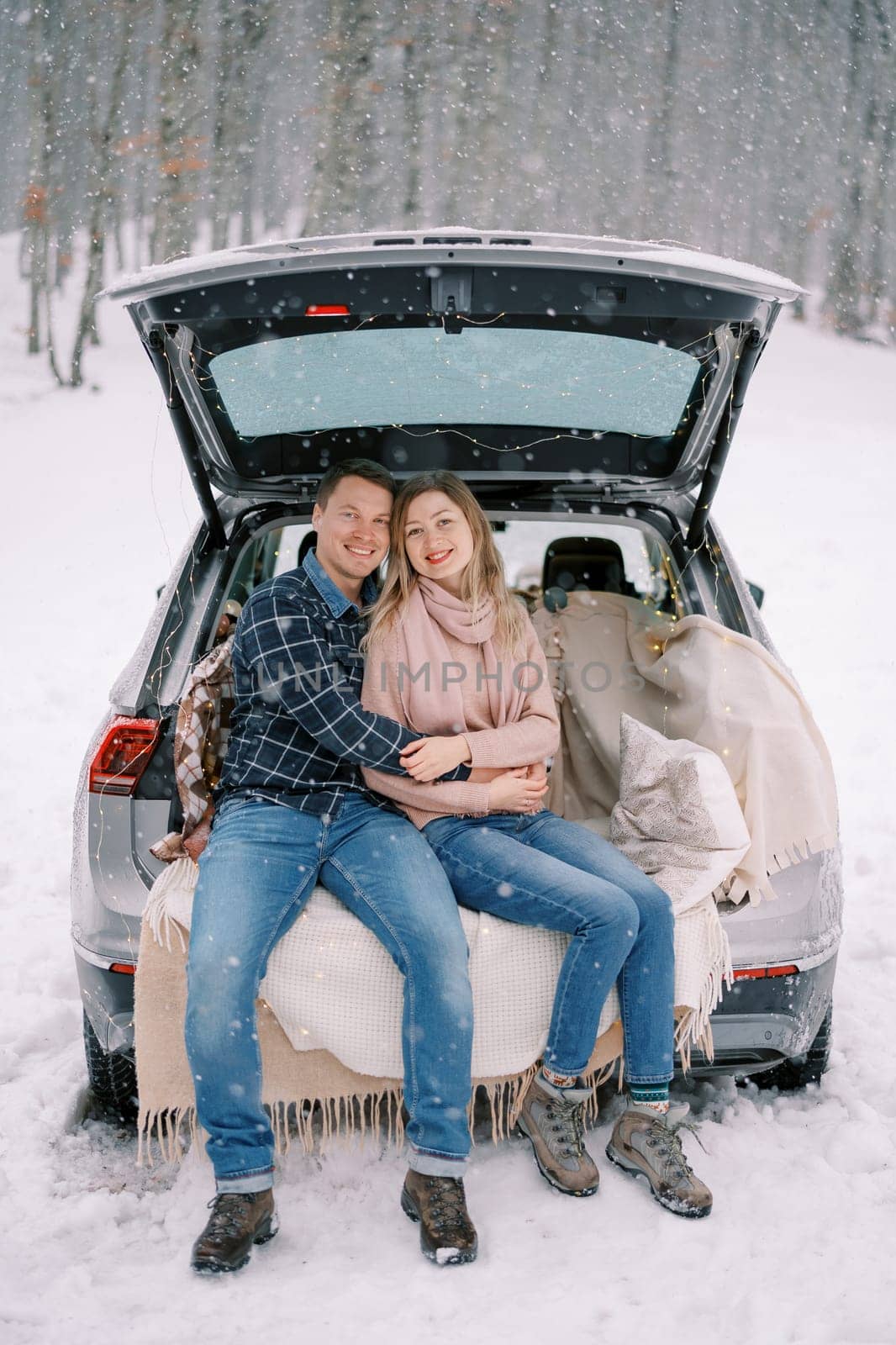 Smiling man and woman sit hugging in car trunk on blankets under snowfall in forest by Nadtochiy