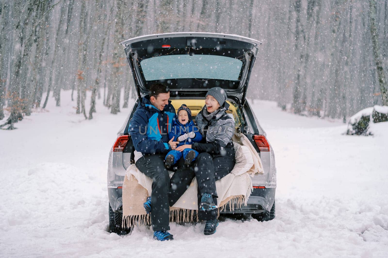 Laughing mom, dad and little girl are sitting on a blanket in the trunk of a car under a snowfall in the forest by Nadtochiy