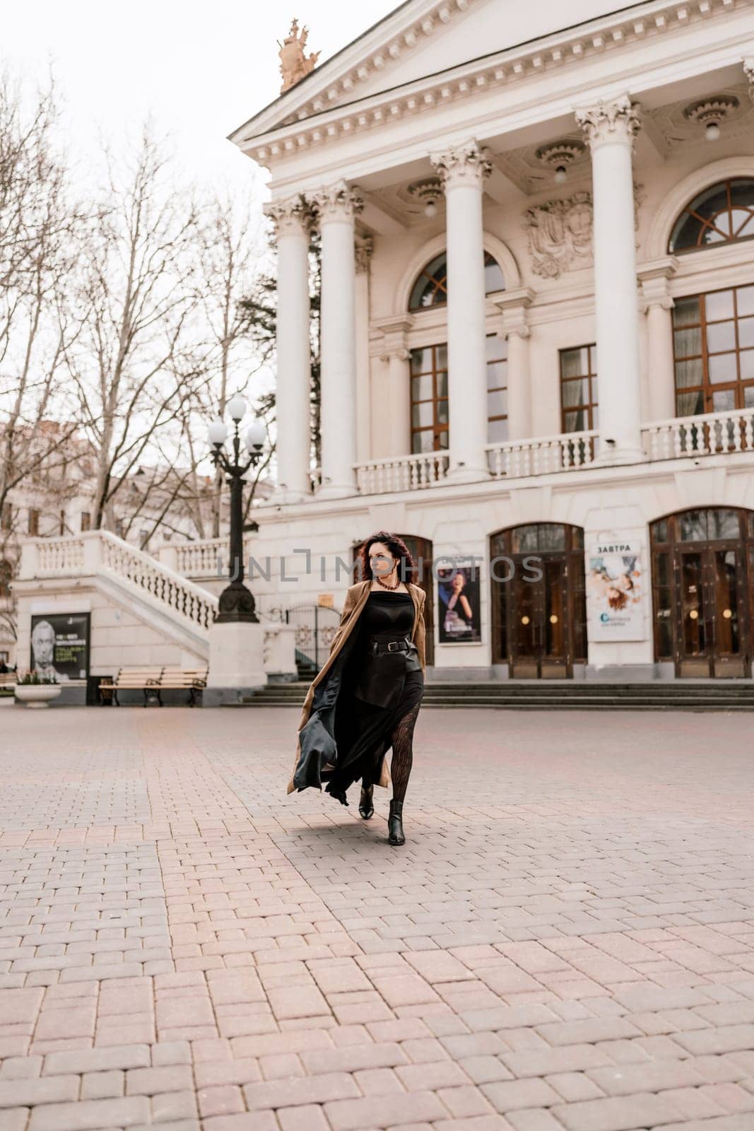 Woman street lifestyle. Image of stylish woman walking through European city on sunny day. Pretty woman with dark flowing hair, dressed in a beige raincoat and black, walks along the building