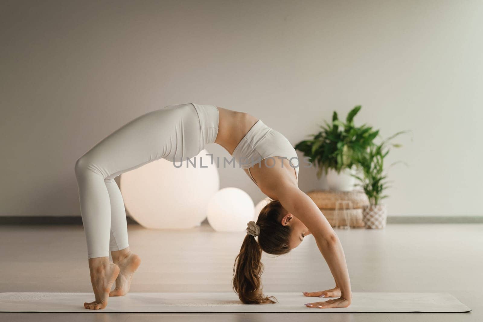 A girl in white clothes does yoga standing on the bridge on a mat indoors.
