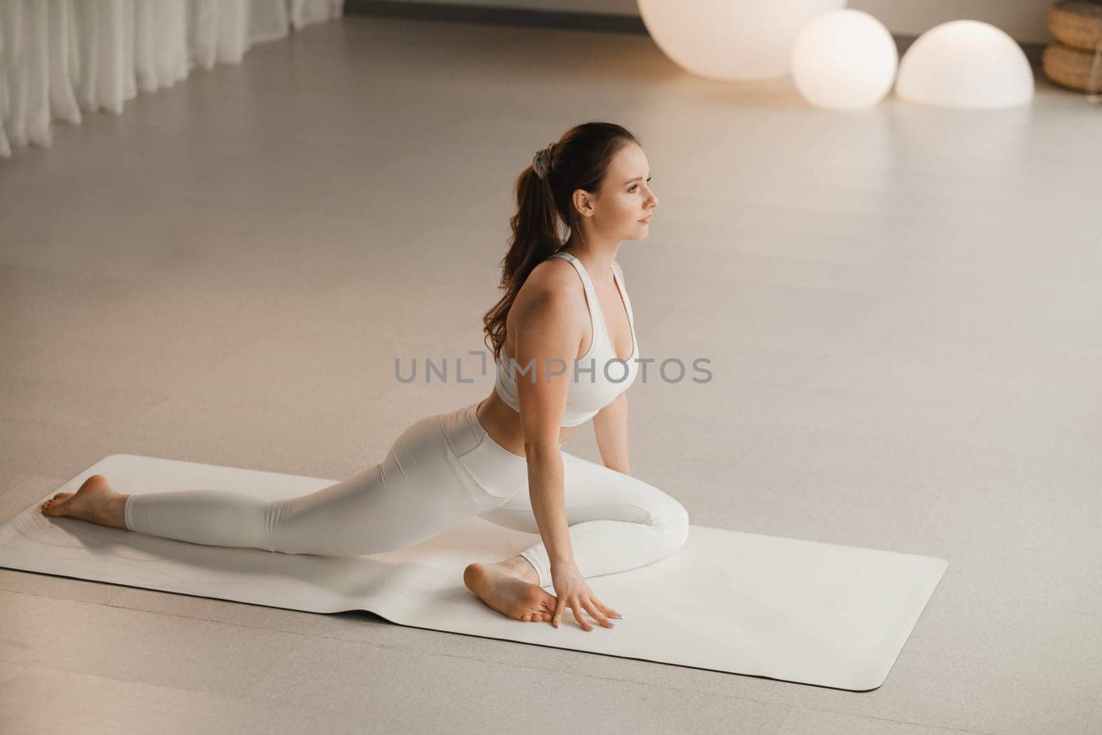 A girl in white clothes does yoga on a mat indoors by Lobachad