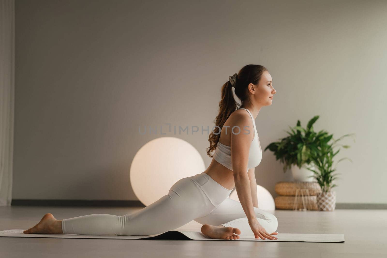 A girl in white clothes does yoga on a mat indoors by Lobachad