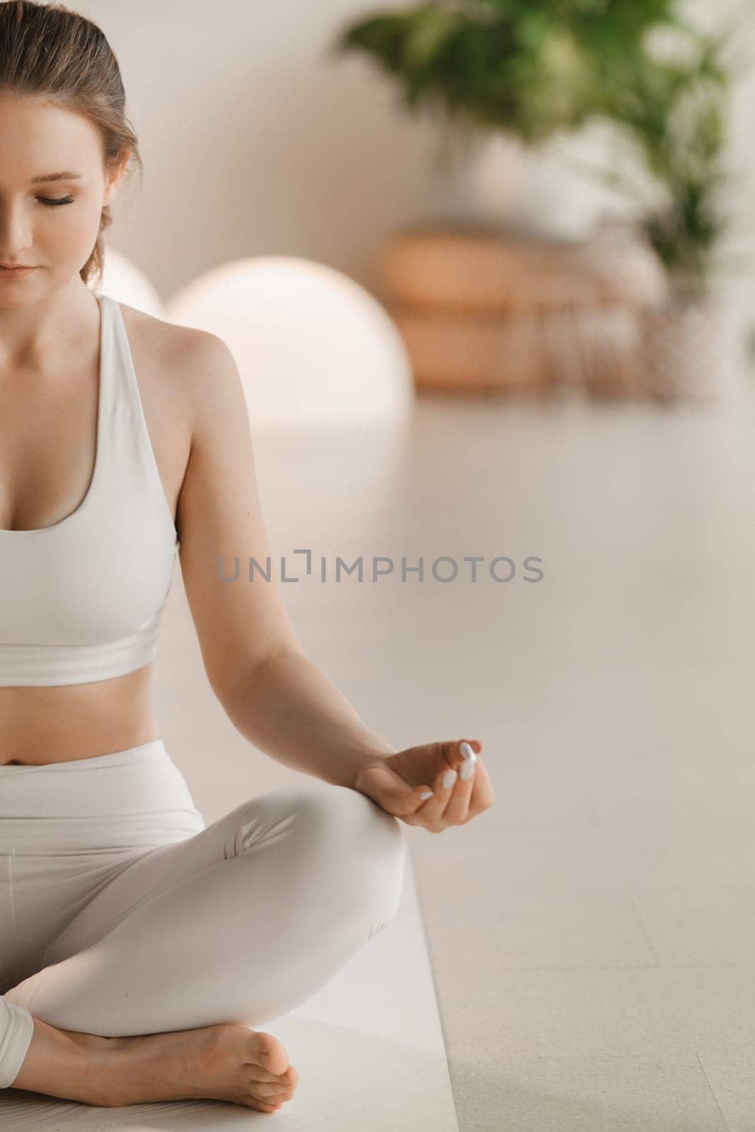 Portrait of a girl in white clothes sitting in a lotus position on a mat at an indoor yoga class by Lobachad