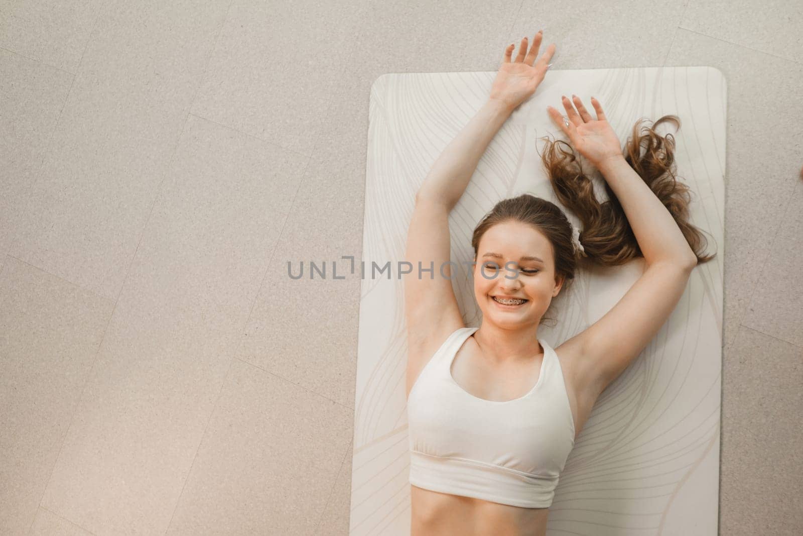 A girl in white clothes lies on a rug and laughs before yoga classes indoors by Lobachad