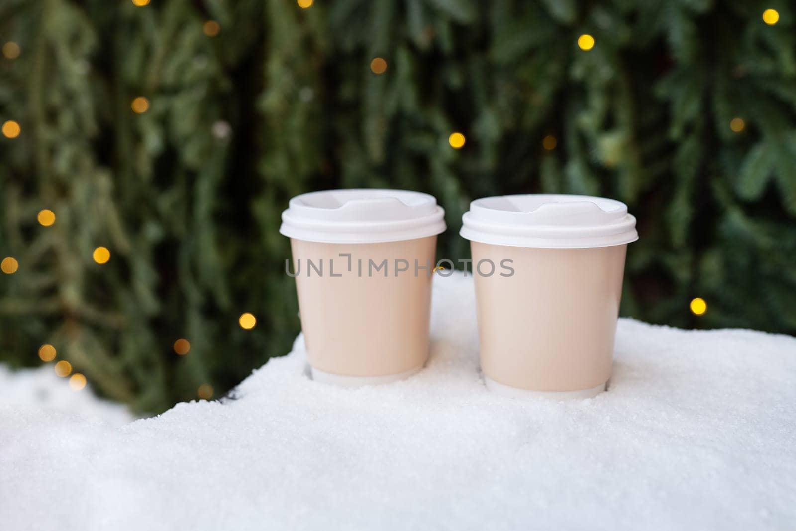 Two plastic cups stand on the snow against the backdrop of a green wall of a Christmas tree and bokeh from a garland. Coffee to go, a walk in the fresh air. by sfinks