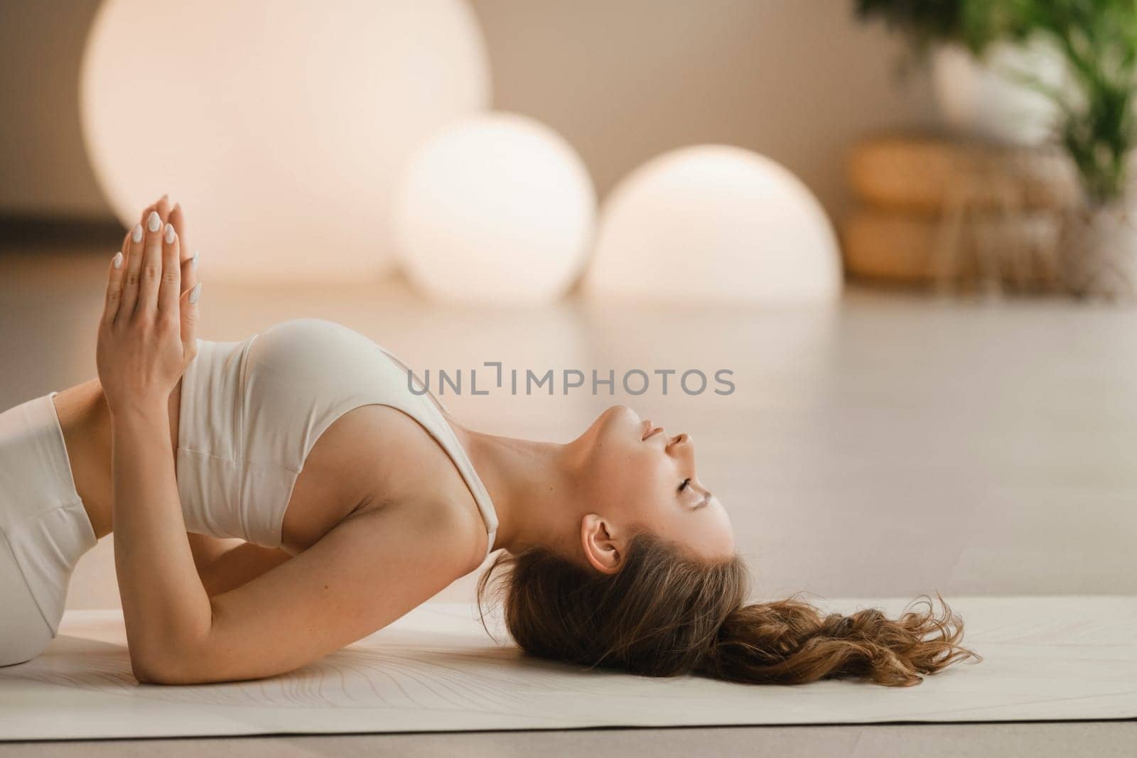 A girl in white clothes does yoga lying on a rug indoors by Lobachad