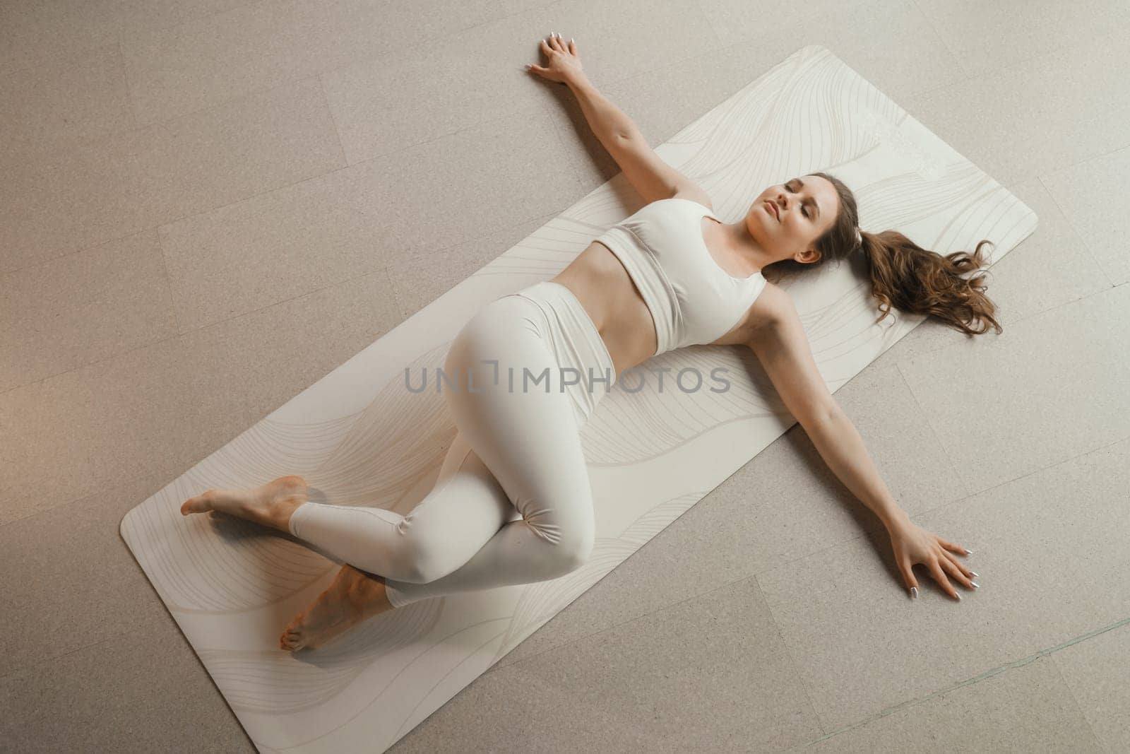 A girl in white clothes does yoga lying on a rug indoors.