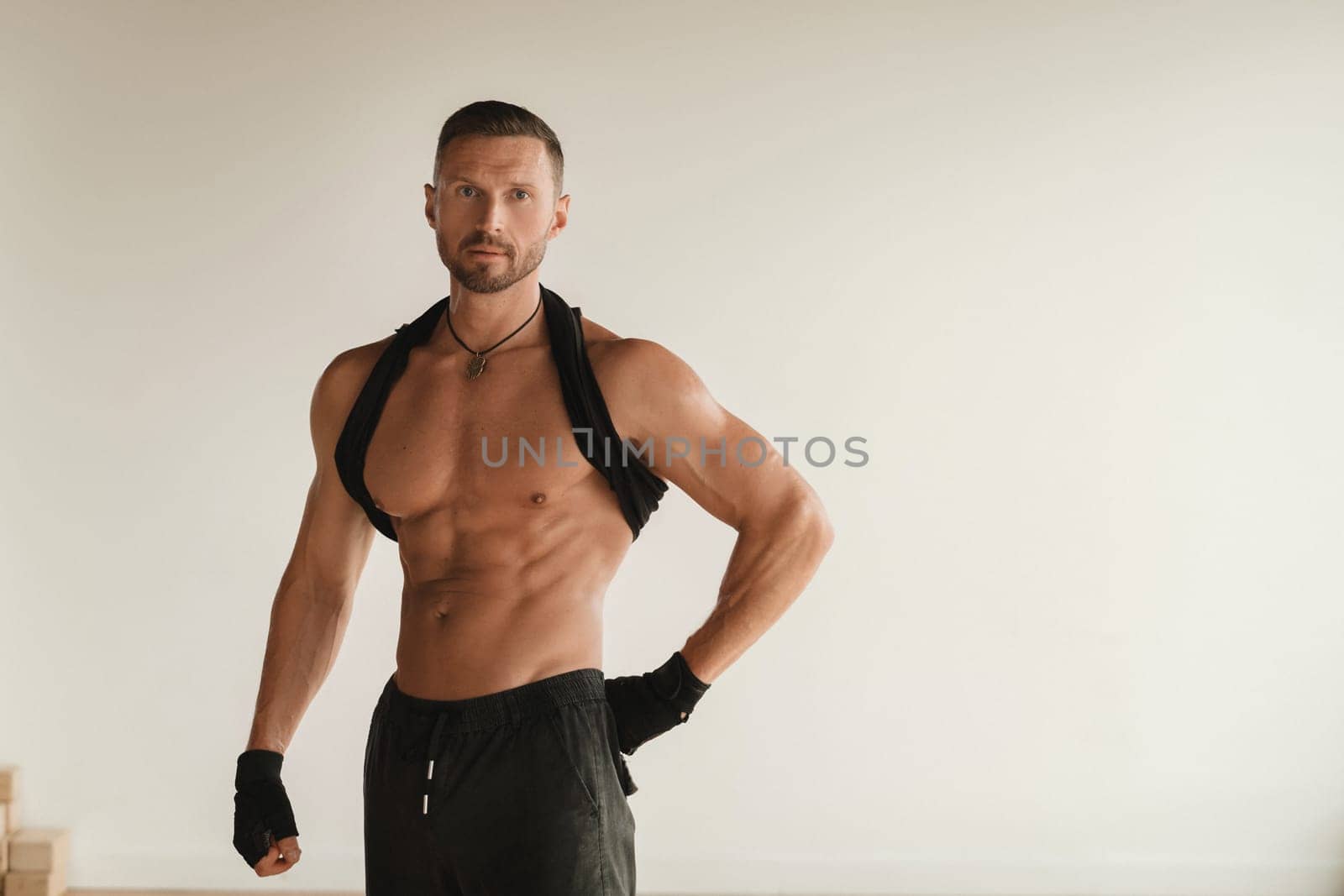 An adult muscular man with a naked torso stands in a room on a light background.