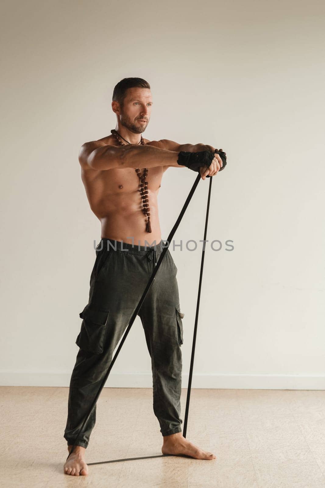 A man with a naked torso is engaged in strength fitness using a rubber loop indoors.