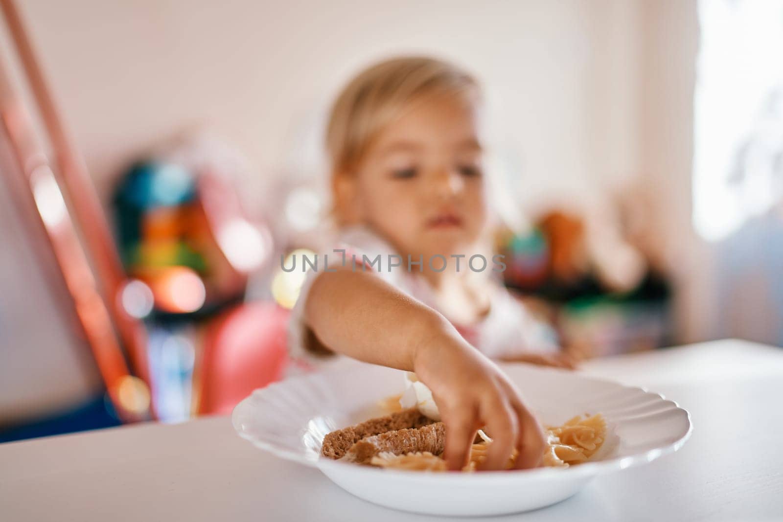Little girl sits at the table and takes out pasta from a plate with her hands by Nadtochiy